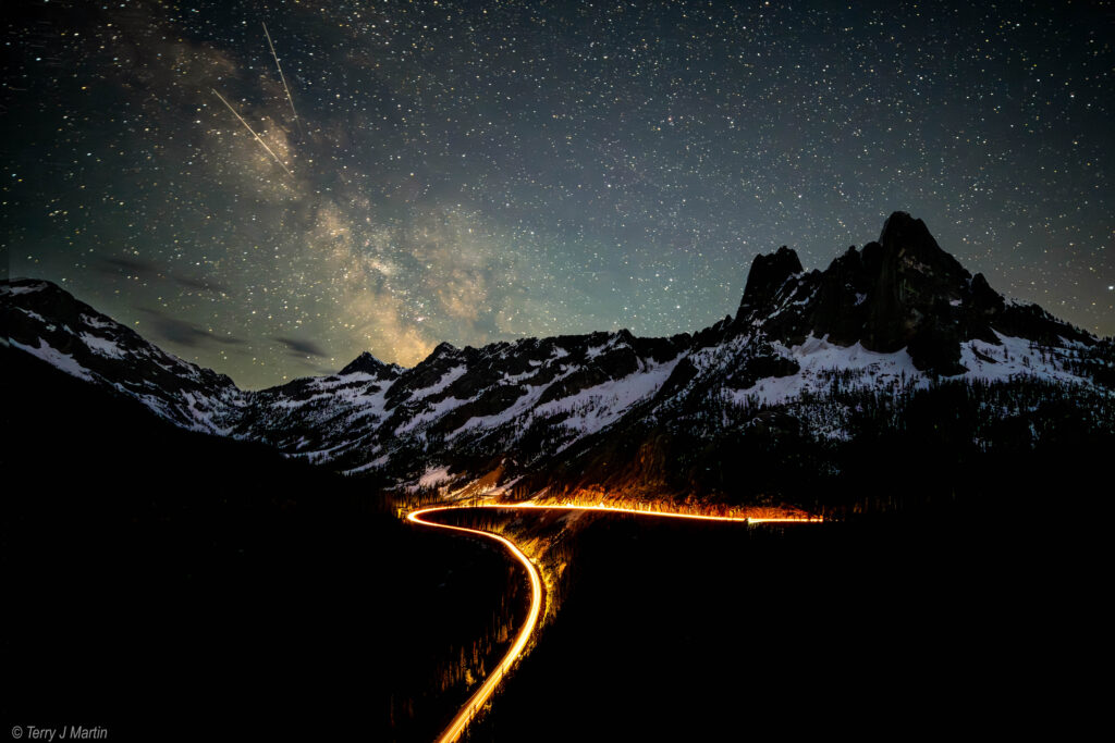A long exposure photograph depicting a firey trail of car headlights in the Northern Cascades park, under the Milky Way in the night sky